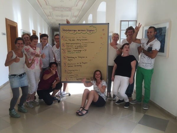 Nach dem Abschluss eines Coachings für Unternehmen steht eine Gruppe zusammen um eine Metaplan Wand und lacht erfolgreich ins Bild.